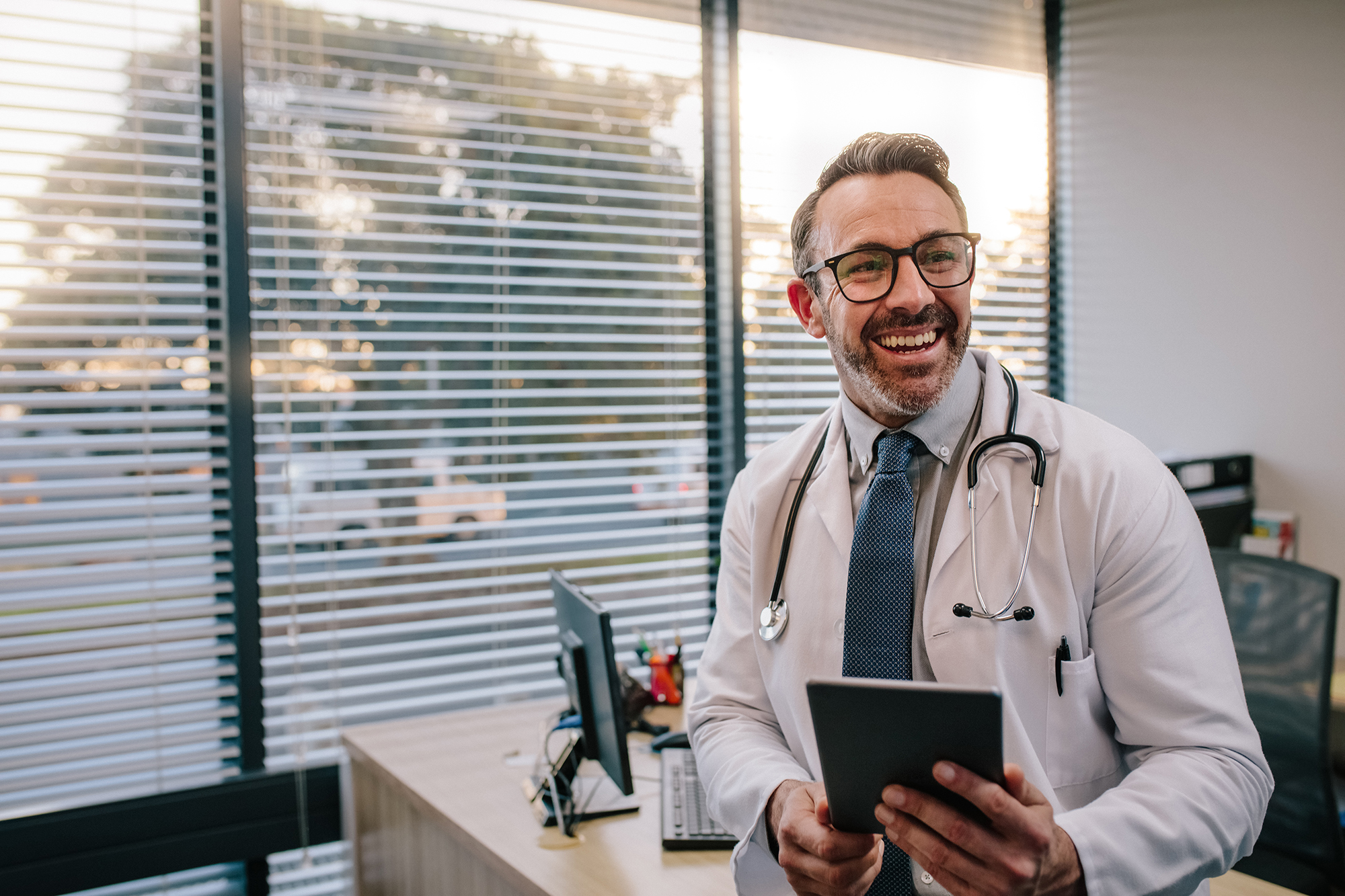 Smiling mature male doctor with digital tablet in his office. Friendly medical professional with tablet computer in clinic.